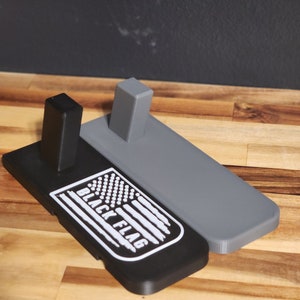 Puede incluir: Un soporte de exhibici&oacute;n impreso en 3D negro y gris con un logotipo "BLACK FLAG" blanco y un dise&ntilde;o de bandera estadounidense.