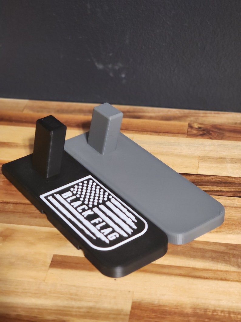 Puede incluir: Un soporte de exhibici&oacute;n impreso en 3D negro y gris con un logotipo "BLACK FLAG" blanco y un dise&ntilde;o de bandera estadounidense.