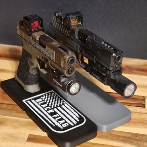 Puede incluir: Dos pistolas negras con miras de punto rojo y linternas montadas en un soporte negro y gris con las palabras "Black Flag" y un dise&ntilde;o de bandera estadounidense.