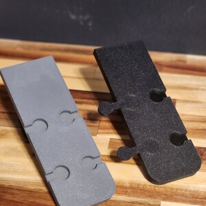 Puede incluir: Dos soportes de pared de pl&aacute;stico, uno gris y otro negro, con m&uacute;ltiples agujeros para colgar objetos. Los soportes est&aacute;n dise&ntilde;ados para ser fijados a una pared.