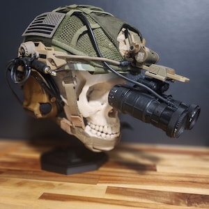 Könnte beinhalten: Taktischer Helm mit Nachtsichtgerät, montiert auf einem Totenkopf. Der Helm hat ein Tarnmuster, einen amerikanischen Flaggenaufnäher und verschiedene Anbauteile. Die Nachtsichtgeräte sind schwarz, das Thema ist militärisch.