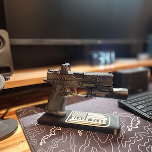 May include: A black and grey handgun with a red dot sight and a flashlight attached to the rail. The gun is mounted on a black stand with a white "BLACK FLAG" logo and an American flag design.