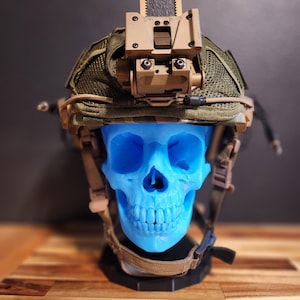 Puede incluir: Un casco militar de color beige y verde con una cubierta de malla y un accesorio de color beige en la parte superior. El casco está sobre un cráneo de plástico azul.