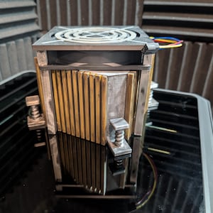 以下が含まれることがあります： 黒いファン、銀色の金属側面、金色のフィンを備えたコンピューターCPUクーラー。 クーラーには2つの銀色のクランプと、側面から伸びるいくつかの色のワイヤーがあります。 オブジェクトは反射する黒い表面にあります。