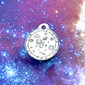 May include: A round, silver-colored pet tag with a space-themed design. The tag features a cartoon cat, planets, stars, a UFO, and a shooting star. The tag has a small hole at the top for attaching to a collar.