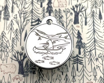 May include: Silver round tag with a bear in a canoe paddling down a river with three fish swimming below. The tag has a hole at the top for attaching to a key chain or dog collar.