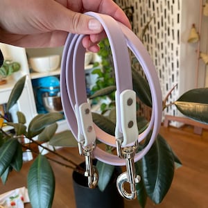 May include: A lavender-colored leash made of a smooth, flexible material. The leash features white hardware connectors that secure the strap to two silver-toned metal swivel snap hooks. The design is simple, with a continuous loop of material.