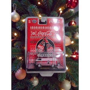 May include: A 1960 VW Delivery Van toy in a clear plastic case, featuring a red and white Coca-Cola design. The packaging has a red background with Coca-Cola branding and snowflake patterns. The toy is displayed against a Christmas tree background.