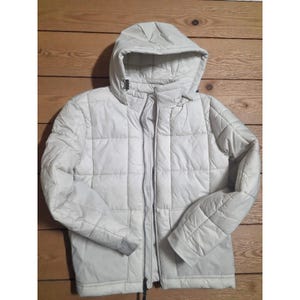 May include: A light-colored, quilted jacket with a hood. The jacket has a full-length zipper and a quilted pattern. The sleeves are long, and the jacket appears to be made of a lightweight material. The jacket is designed for cold weather.