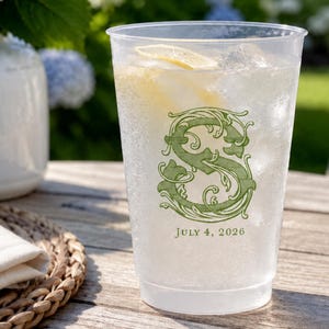 May include: A clear plastic cup filled with ice and a lemon slice, featuring a green decorative letter S and the printed text JULY 4, 2026. The cup has a tapered cylindrical shape and is positioned on a wooden surface.