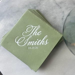 May include: A stack of square sage green paper napkins featuring white printed text that reads The Smiths and the date 04.20.26 in a cursive script font. The napkins are layered and have a smooth, matte finish with clean, sharp edges.