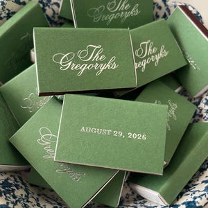 May include: A collection of rectangular matchboxes with a matte sage green finish. Each box features the text The Gregoryks in elegant silver script or the date AUGUST 29, 2026, printed in a clean, sans-serif silver font on the front cover.