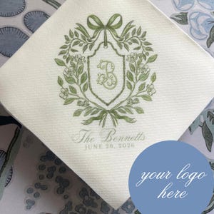 May include: A square white paper napkin featuring a green botanical wreath illustration surrounding a central shield with the letter B. Below the shield, the text reads The Bennetts and JUNE 26, 2026. A blue circle with the text your logo here is present.