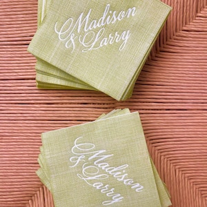 Pu&ograve; includere: Pile di tovaglioli quadrati verde chiaro con i nomi "Madison & Larry" in elegante scrittura bianca. I tovaglioli hanno un aspetto strutturato e sono disposti su una superficie intrecciata marrone chiaro. Ideale per un matrimonio o un evento speciale.