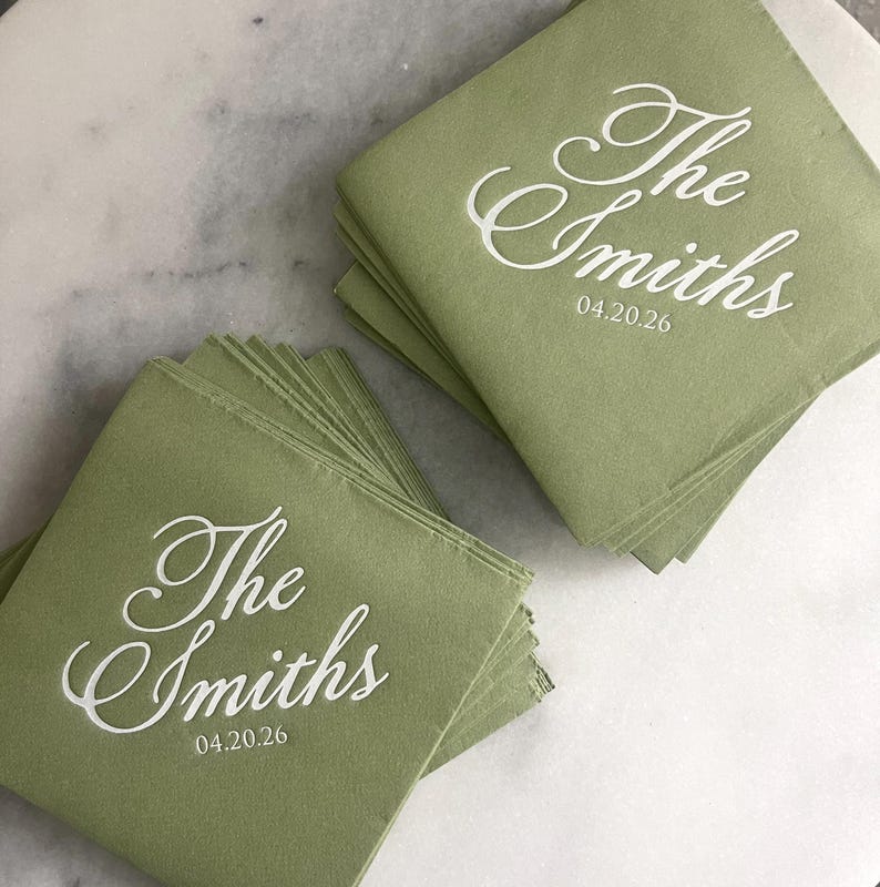 May include: A stack of square sage green paper napkins featuring white printed text that reads The Smiths and the date 04.20.26 in a cursive script font. The napkins are arranged in two separate piles, showcasing the custom lettering clearly.