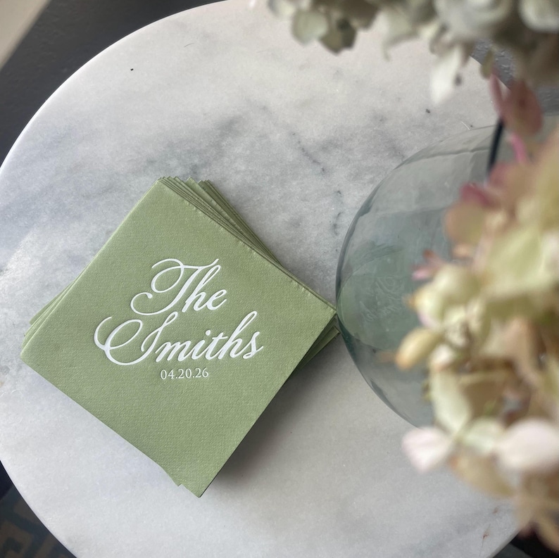 May include: A stack of square sage green paper napkins featuring white printed text that reads The Smiths and the date 04.20.26 in a cursive script font. The napkins are layered and have a smooth, matte finish with clean, sharp edges.