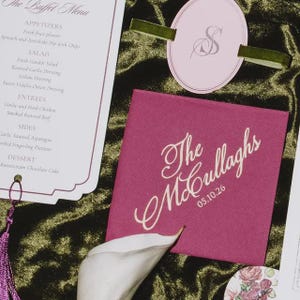 May include: A white buffet menu card with black text, a pink circular tag with a silver monogram and green velvet ribbon, and a square magenta card featuring the white script text The McCullaghs and the date 05.10.26, alongside a white calla lily.
