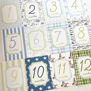 May include: A set of twelve baby milestone cards, each featuring a number from 1 to 12, with the word "months" below. The cards have various patterned backgrounds in blue, green, and white, with decorative frames.