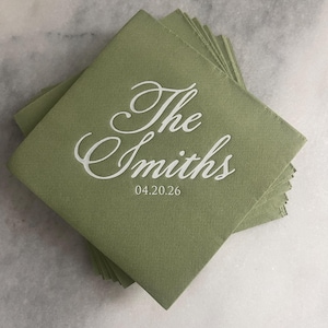 May include: A stack of square sage green paper napkins featuring white printed text that reads The Smiths in a cursive script font, with the date 04.20.26 printed in a smaller, clean sans-serif font directly underneath the family name.