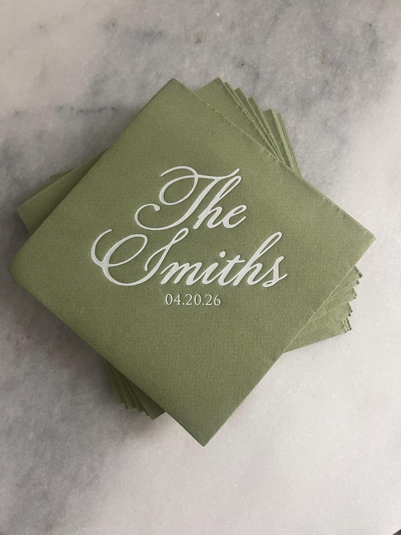 May include: A stack of square sage green paper napkins featuring white printed text that reads The Smiths in a cursive script font, with the date 04.20.26 printed in a smaller, clean sans-serif font directly underneath the family name.