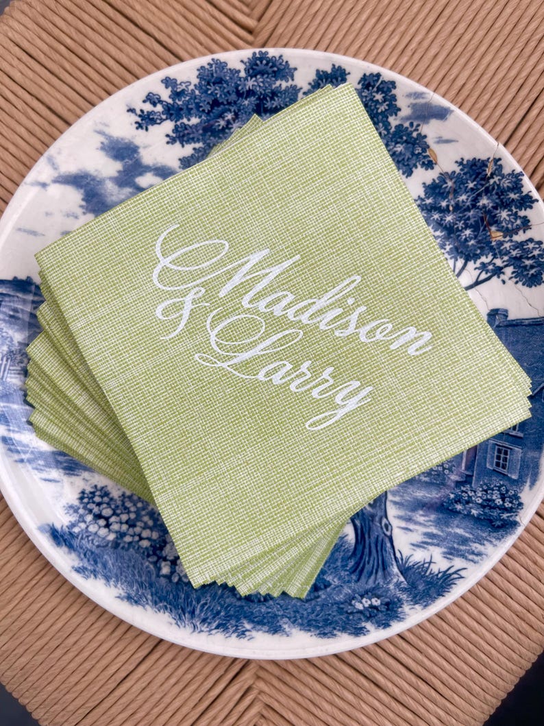 Pu&ograve; includere: Una pila di tovaglioli di lino verde chiaro con i nomi "Madison & Larry" ricamati in corsivo bianco. I tovaglioli poggiano su un piatto decorativo blu e bianco con un disegno paesaggistico.