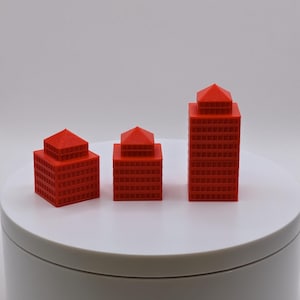 May include: Three red miniature building models of varying sizes. Each model has a square base and a pyramidal roof. The buildings are arranged on a white circular platform against a plain white background. The models are likely 3D printed.