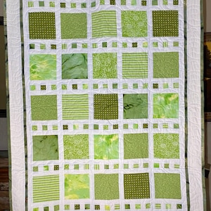 May include: A green and white quilt with a patchwork design. The quilt is made up of squares of different green fabrics, each with a different pattern. The quilt is bordered with a white fabric and a green fabric.