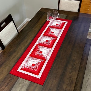 May include: A red and white quilted table runner with a geometric pattern. The runner is on a dark wood table with two chairs.