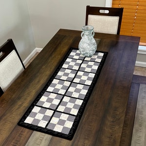 May include: A black and white checkered table runner with a decorative border. The runner is on a dark brown wooden table with a clear glass vase in the center.