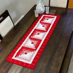 May include: A red and white quilted table runner with a geometric pattern. The runner is on a dark wood table with two chairs and a glass vase.
