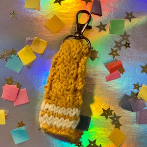 May include: Mustard yellow and white crocheted keychain with a metal clasp and star charm. The keychain is made of a thick, textured yarn and features a striped design at the bottom. The background is a holographic surface with star and square confetti.