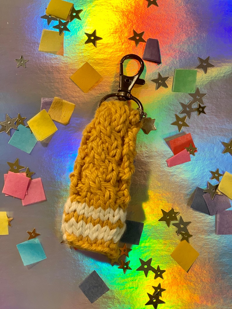 May include: Mustard yellow and white crocheted keychain with a metal clasp and star charm. The keychain is made of a thick, textured yarn and features a striped design at the bottom. The background is a holographic surface with star and square confetti.