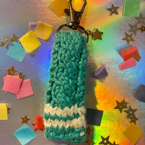 May include: A teal and white knitted keychain with a silver clasp and star charm. The keychain is made of a teal yarn with a braided pattern and white stripes near the bottom. The background is a shiny, iridescent surface with colorful confetti and star-shaped decorations.