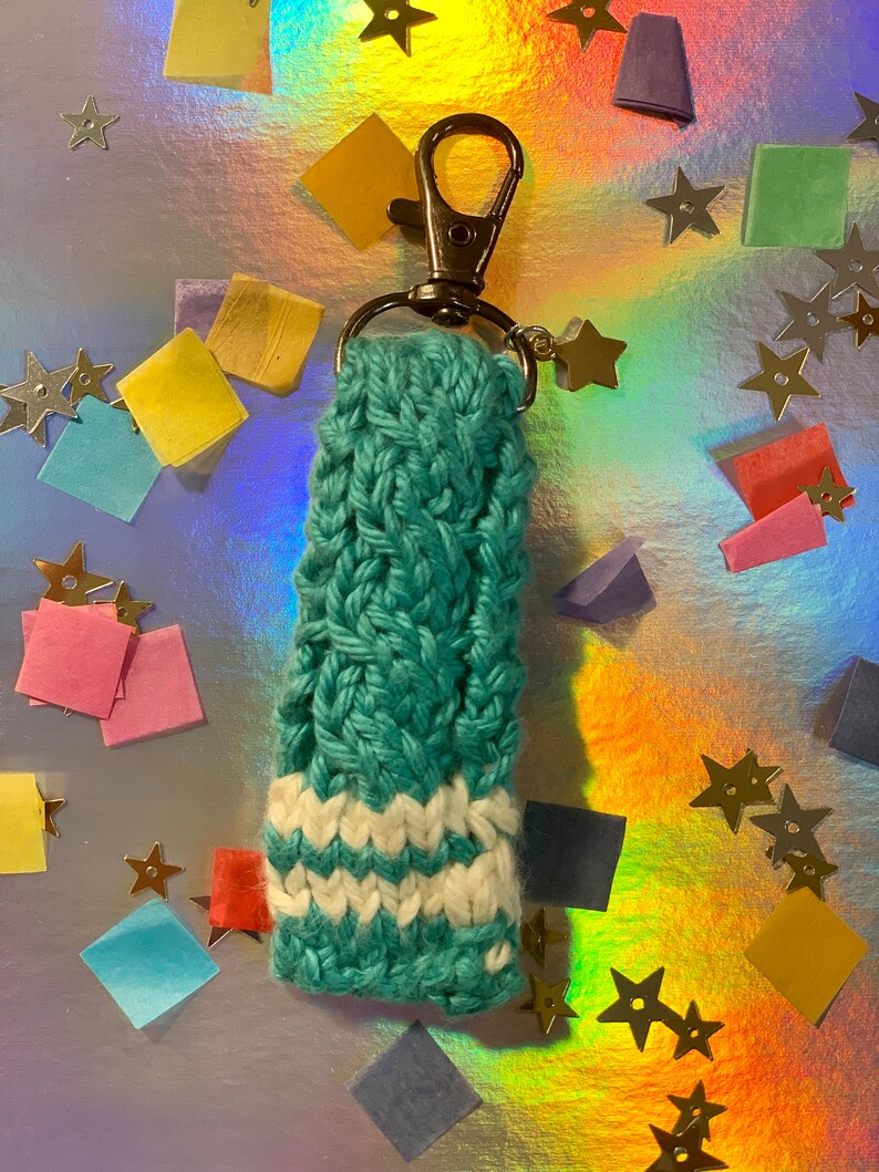 May include: A teal and white knitted keychain with a silver clasp and star charm. The keychain is made of a teal yarn with a braided pattern and white stripes near the bottom. The background is a shiny, iridescent surface with colorful confetti and star-shaped decorations.