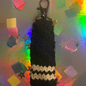 May include: A black and white knitted keychain accessory with a silver clasp. The accessory features a black body with a white zig-zag pattern near the bottom. The background is a shiny, iridescent surface with colorful confetti and star-shaped decorations.