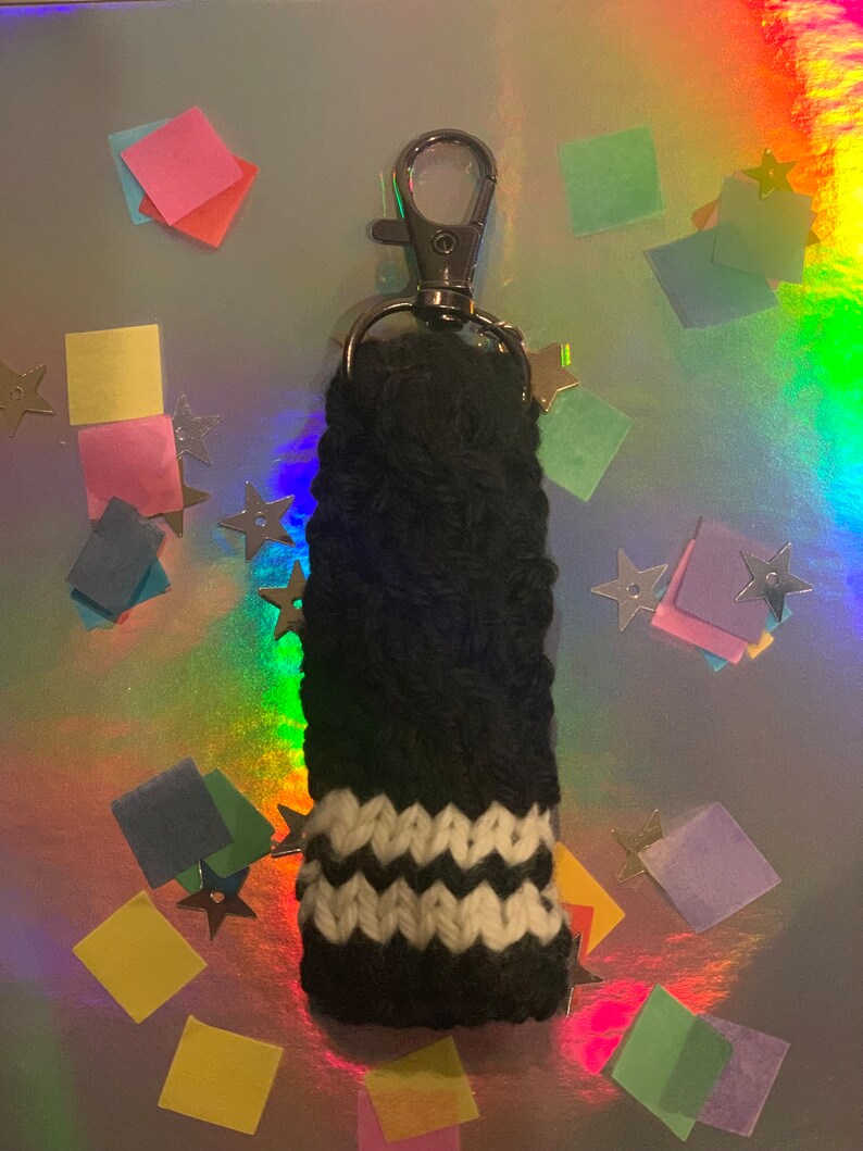May include: A black and white knitted keychain accessory with a silver clasp. The accessory features a black body with a white zig-zag pattern near the bottom. The background is a shiny, iridescent surface with colorful confetti and star-shaped decorations.