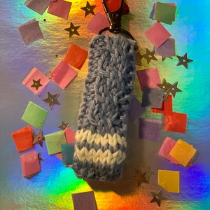 May include: A handmade, blue and white knitted keychain holder with a silver clasp. The holder features a textured, woven design and a white chevron pattern at the base. The background is a shimmering, iridescent surface with scattered confetti and star-shaped decorations.