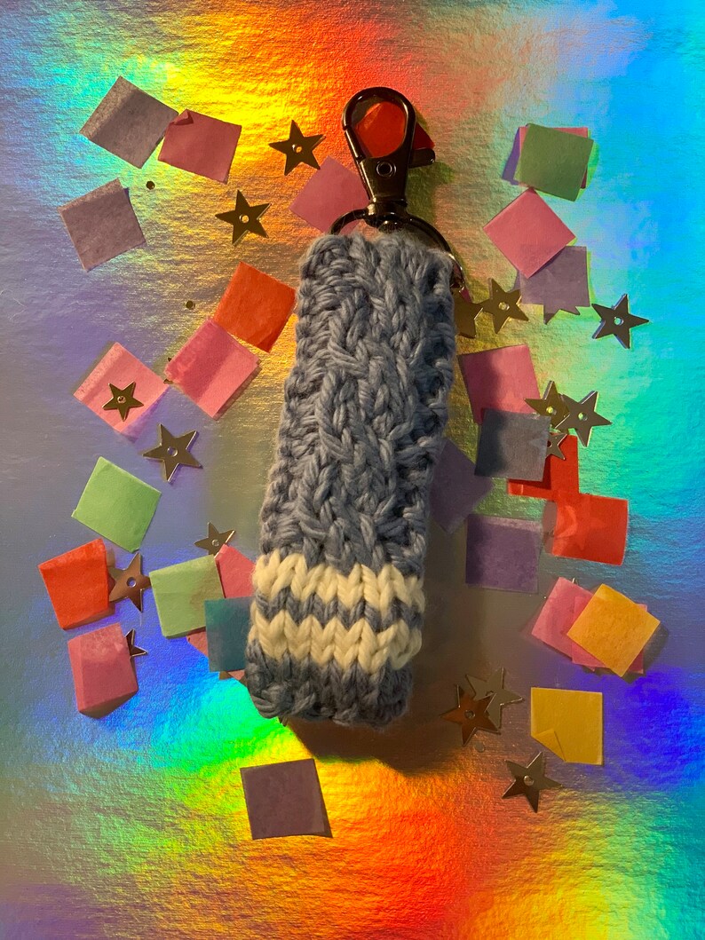 May include: A handmade, blue and white knitted keychain holder with a silver clasp. The holder features a textured, woven design and a white chevron pattern at the base. The background is a shimmering, iridescent surface with scattered confetti and star-shaped decorations.