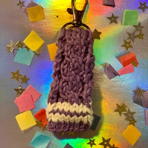 May include: A handmade, purple knitted keychain holder with a white and purple striped design at the bottom. The keychain has a bronze-colored clasp. The background is a shiny, iridescent surface with gold star confetti and colorful paper squares.