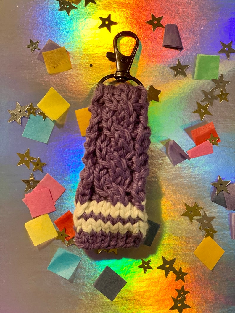 May include: A handmade, purple knitted keychain holder with a white and purple striped design at the bottom. The keychain has a bronze-colored clasp. The background is a shiny, iridescent surface with gold star confetti and colorful paper squares.
