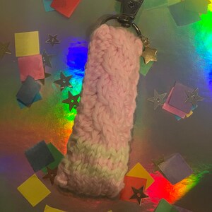 May include: A handmade, pink and white knitted keychain holder with a silver clasp and a gold star charm. The knitted portion has a braided design. The background is a holographic surface with colorful confetti and star-shaped decorations.