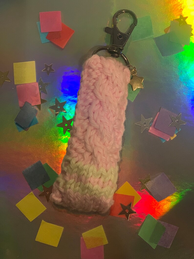 May include: A handmade, pink and white knitted keychain holder with a silver clasp and a gold star charm. The knitted portion has a braided design. The background is a holographic surface with colorful confetti and star-shaped decorations.