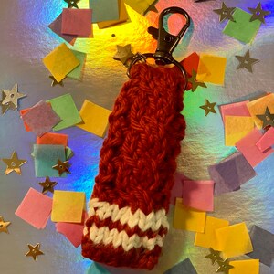 May include: A handmade, red-orange knitted keychain sleeve with a white and off-white striped pattern at the bottom. The sleeve is attached to a silver-colored metal clasp and a small star charm. The background is a shiny, iridescent surface with colorful confetti.