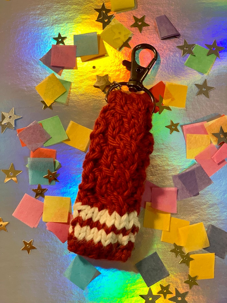 May include: A handmade, red-orange knitted keychain sleeve with a white and off-white striped pattern at the bottom. The sleeve is attached to a silver-colored metal clasp and a small star charm. The background is a shiny, iridescent surface with colorful confetti.