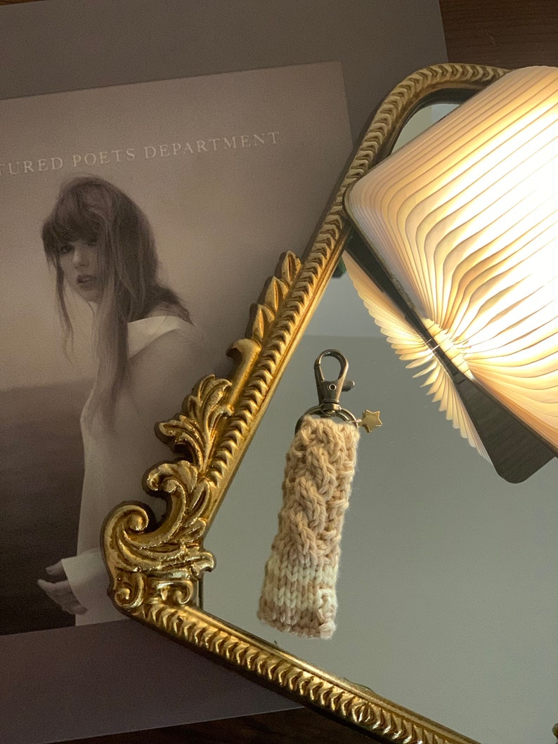 May include: A handmade, knitted keychain in cream and tan hues, with a gold-colored star charm. The keychain is displayed in front of a gold-framed mirror, with a book and a book cover visible in the background. The book cover reads "The Tortured Poets Department."