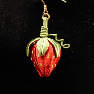 Op de afbeelding: Een enkele handgemaakte aardbeienooring. De rode stoffen bes heeft goudkleurige accenten en groene bladeren. De oorbel hangt aan een goudkleurige haak. De achtergrond is zwart.