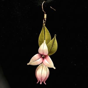 Puede incluir: Un solo pendiente floral con un gancho dorado. El pendiente presenta un grupo de pétalos rosas y blancos, con tres hojas verdes encima. El pendiente está sobre un fondo negro.