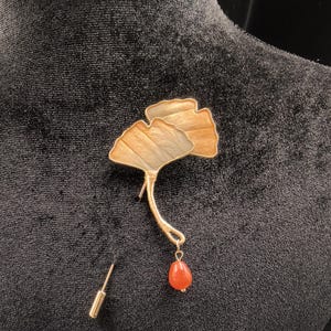 Könnte beinhalten: Eine goldfarbene Brosche in Form eines Ginkgoblattes mit einer tropfenförmigen, orangefarbenen Perle. Die Brosche ist auf einem schwarzen Samtstoff befestigt, was ihr detailliertes Design und ihre Eleganz hervorhebt. Die Nadel ist separat.