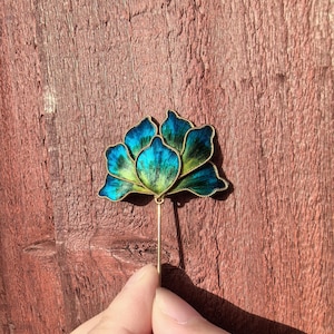 Puede incluir: Un broche floral artesanal con un vibrante diseño de esmalte azul y verde. El broche presenta un contorno de metal dorado y un alfiler largo y delgado. Los pétalos de la flor tienen un efecto degradado, pasando de un azul profundo a un verde más claro.