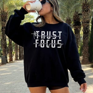 May include: A black long-sleeve sweatshirt featuring white text that reads TRUST THE PROCESS and FOCUS ON YOUR JOURNEY. The garment has a crew neckline and ribbed cuffs, providing a standard fit. The design is centered on the chest area.
